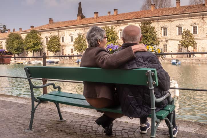 Elderly couple enjoy Lake Garda's scenic view, embracing on bench.