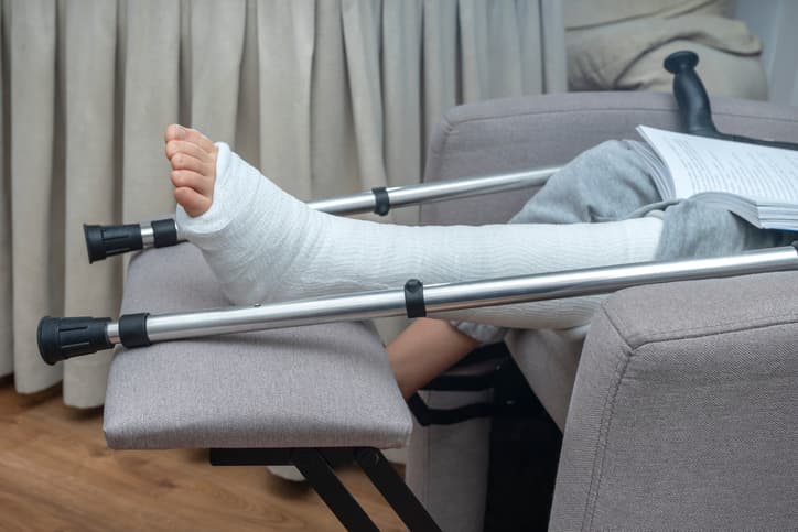 CHILD WITH CASTED BROKEN LEG AND CRUTCHES ON FOLDING ARMCHAIR AND READING A BOOK AT HOME